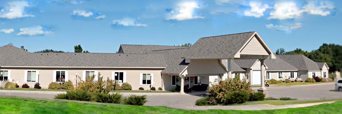 Exterior view of a single-story senior living facility building with beige siding and a covered entrance. The building is surrounded by green grass, bushes, and a paved driveway under a partly cloudy blue sky.