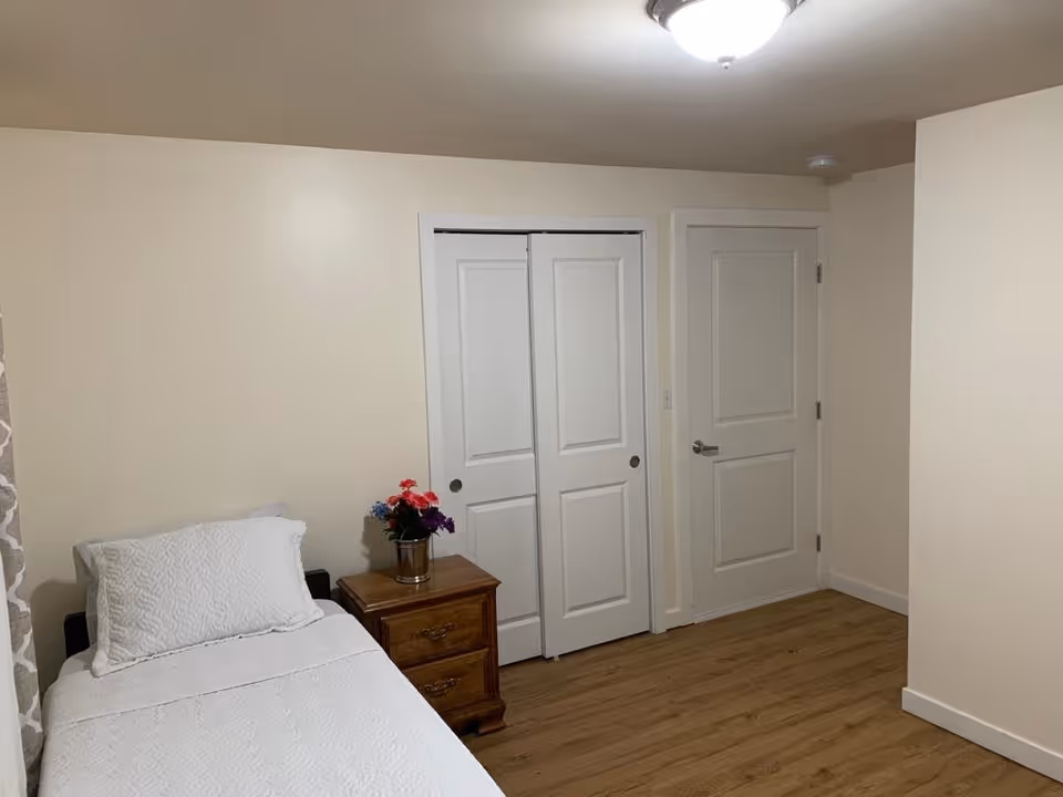 A simple bedroom with a single bed covered in white bedding, a wooden nightstand with a small flower pot containing colorful flowers, light-colored walls, wooden flooring, a closed white door, and a white closet with sliding doors.