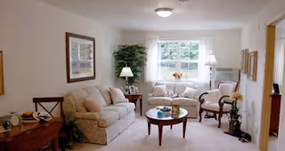 A cozy living room with a beige sofa, two armchairs, a round wooden coffee table with decorative items, a side table with a lamp, framed artwork on the walls, a large window with white curtains, and a potted plant in the corner.