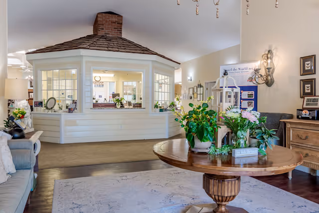 A bright and welcoming interior common area in a senior living facility featuring a central wooden table with green plants and flowers, a cozy seating area with a sofa on the left, and a unique octagonal structure with windows and a shingled roof in the background. The room has warm lighting and decorative wall sconces.