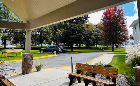 Covered entryway with benches overlooking a driveway, parked cars, trees, and a grassy courtyard.