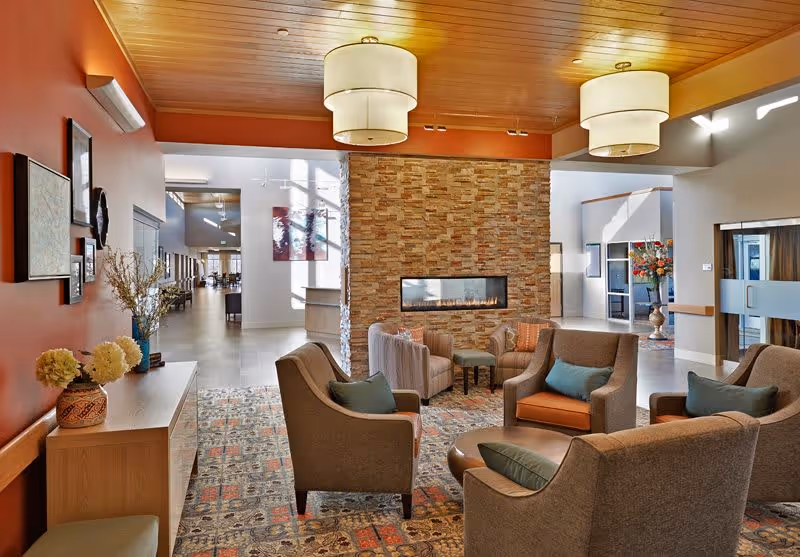Cozy lobby seating area with several armchairs arranged around a low table facing a long gas fireplace set in a stone feature wall under pendant lights.