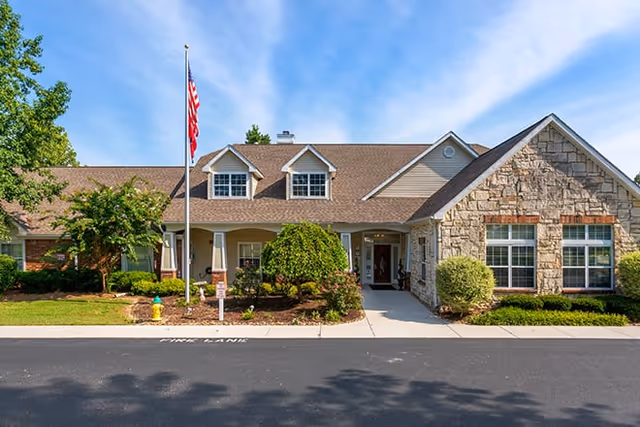Front exterior view of a single-story senior living facility building with a stone facade and beige siding. The building has a brown shingled roof with dormer windows, a covered entrance, and an American flag on a flagpole in front. There are well-maintained shrubs, trees, and a fire hydrant near the sidewalk under a blue sky with some clouds.