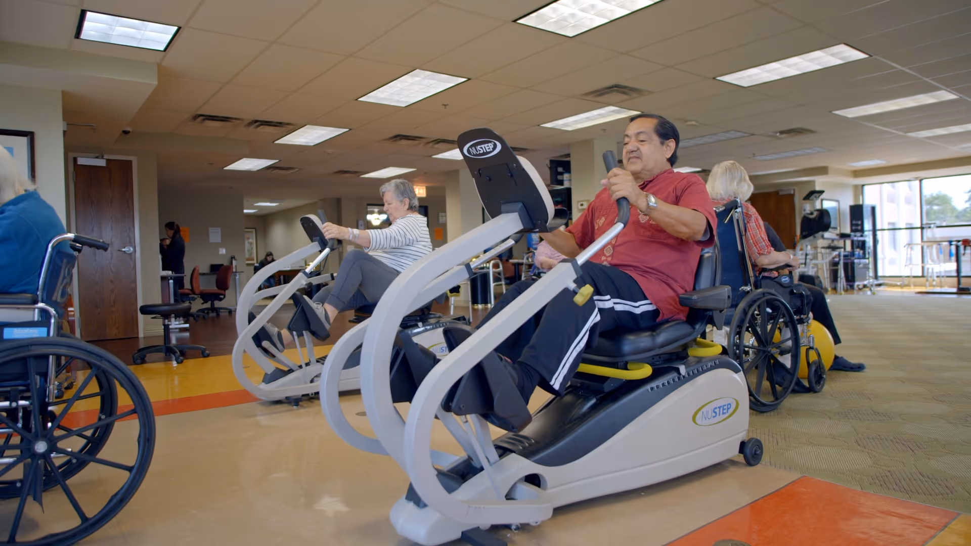 Several elderly individuals exercising on seated elliptical machines in a spacious, well-lit fitness room with large windows and wheelchairs nearby.