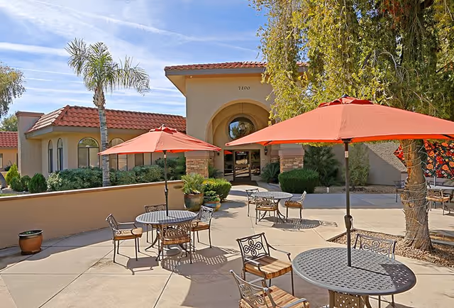 Outdoor patio area at Pueblo Norte Senior Living with round metal tables and chairs, each shaded by large red umbrellas. The patio is surrounded by desert landscaping including palm trees and shrubs, with a beige building featuring a tiled roof and arched entrance in the background under a blue sky.