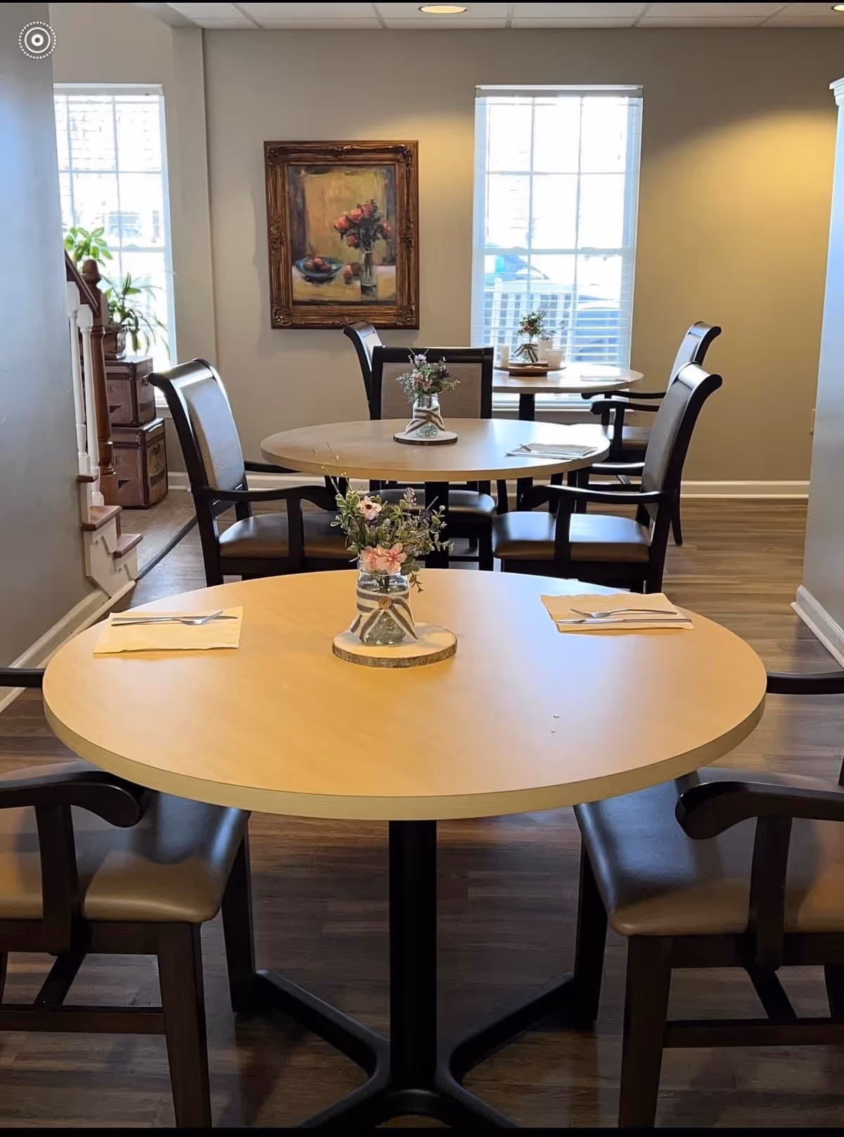 A dining area with round wooden tables and cushioned chairs. Each table has a small floral centerpiece and napkins with utensils. The room has wooden flooring, two large windows letting in natural light, and a framed painting on the wall.