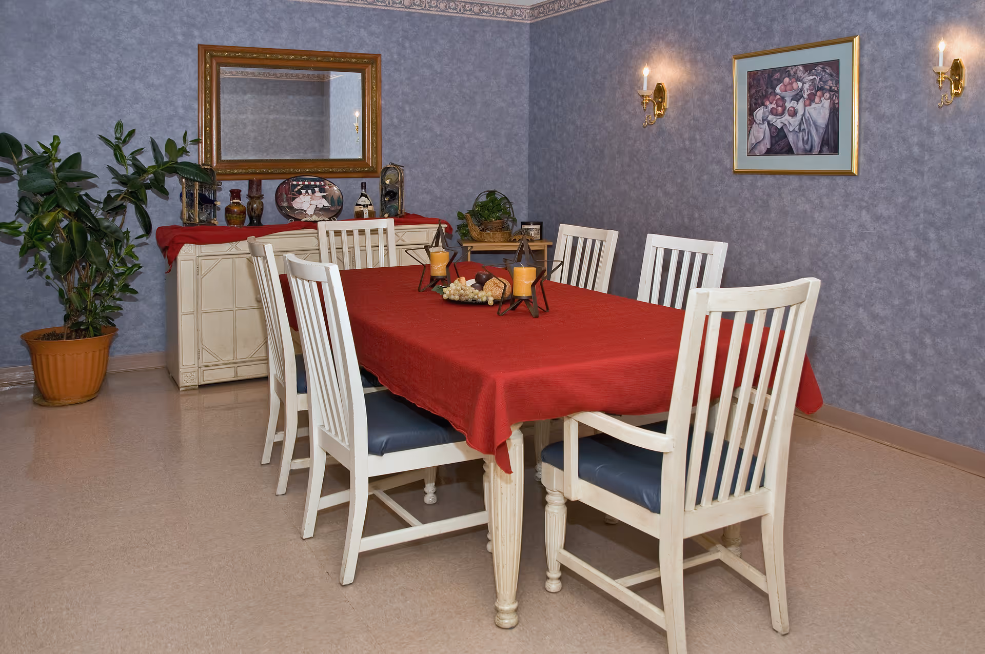 A dining room with a rectangular table covered by a red tablecloth, six white chairs, a sideboard, wall sconces and a potted plant.