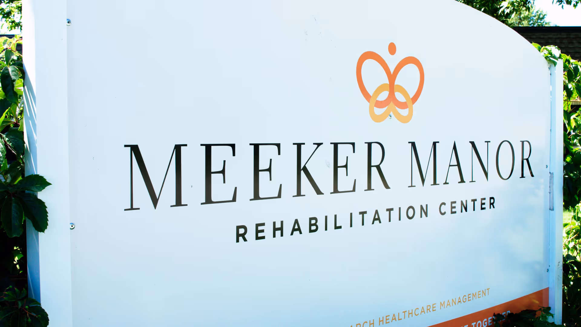 A white outdoor sign for Meeker Manor Rehabilitation Center with a butterfly logo above the name, surrounded by greenery.