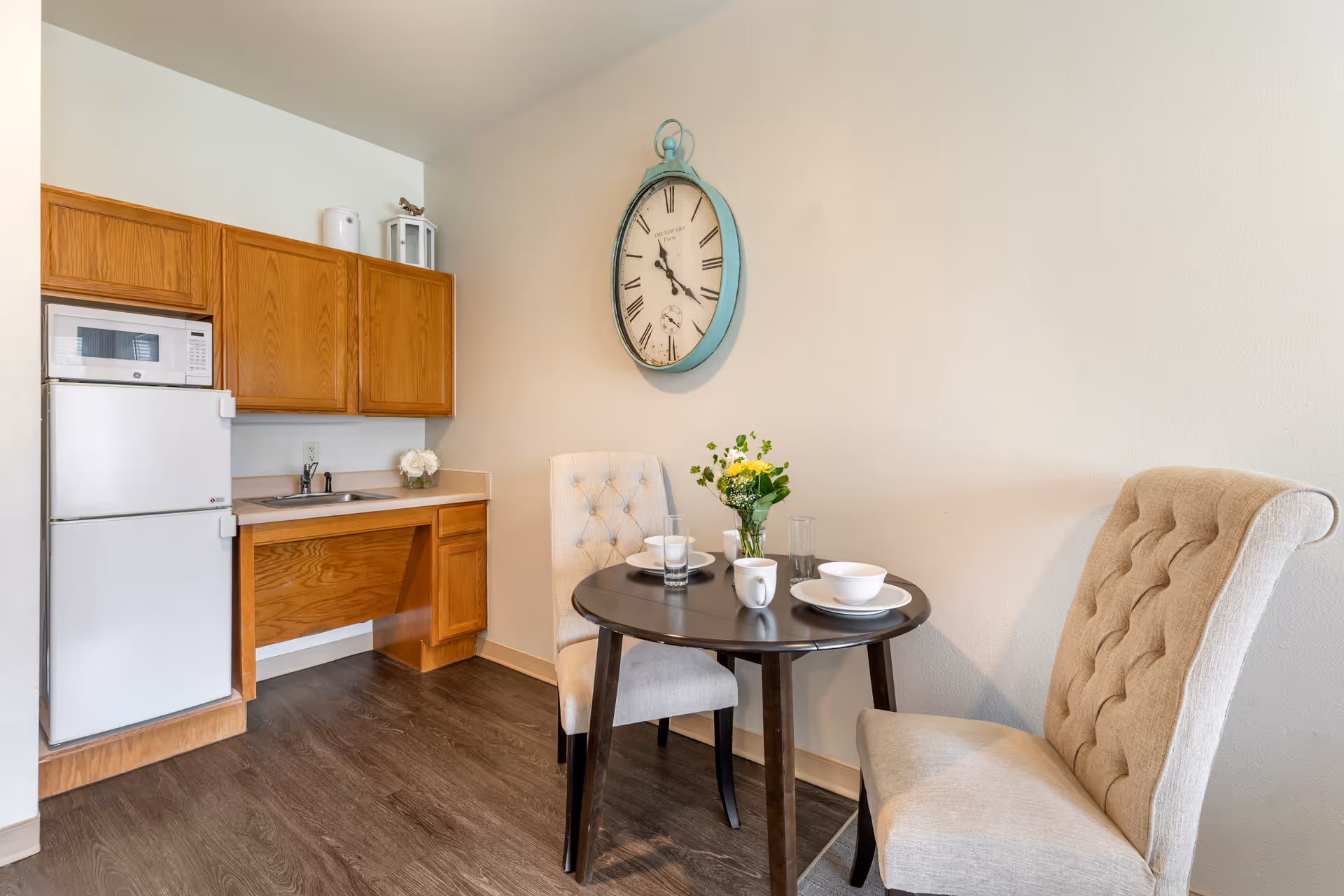 A small dining area with a round dark wooden table set for two, featuring white dishes, cups, and glasses. Two beige upholstered chairs with tufted backs are positioned around the table. On the wall above the table hangs a large vintage-style clock with Roman numerals. To the left is a compact kitchenette with wooden cabinets, a white refrigerator, a microwave, and a small sink. The floor is dark wood, and the walls are light-colored.
