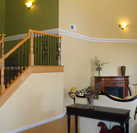 Interior view of a living room area with beige walls, a staircase with wooden handrails and black balusters, a dark wooden table with a vase of flowers, a decorative fireplace, and wall-mounted light fixtures.