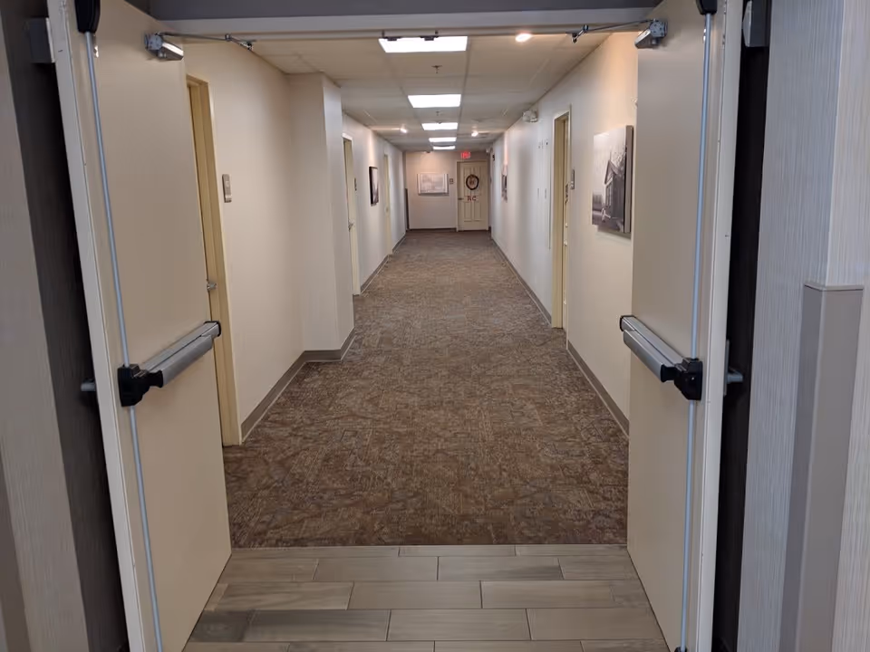 View down a long, carpeted hallway in a supportive living facility with beige walls and several closed doors on both sides. The hallway is lit by ceiling lights and has framed pictures hanging on the walls. Double doors are open in the foreground.