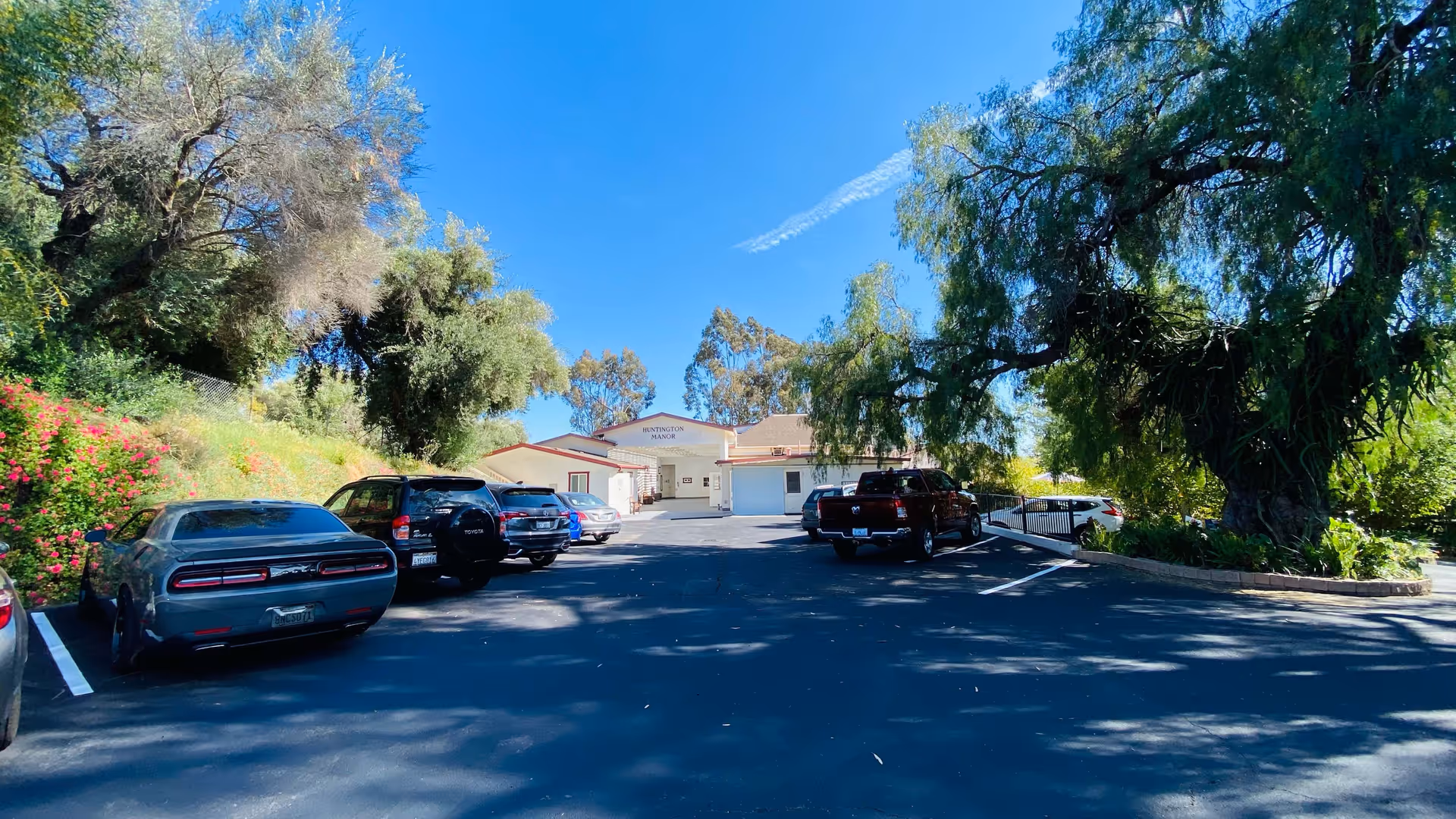 Parking lot with several cars parked on either side, large trees providing shade, and a single-story building in the background with a sign that reads 'Huntington Manor' under a clear blue sky.