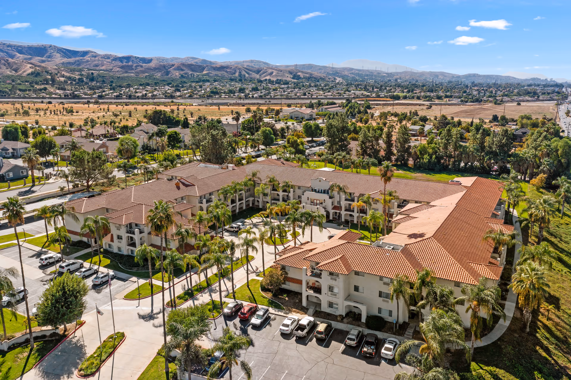 Aerial view of Mission Commons, a senior living community with a large, U-shaped building featuring red tile roofs and surrounded by palm trees and parking lots. The facility is set in a suburban area with hills and residential neighborhoods in the background under a blue sky with scattered clouds.