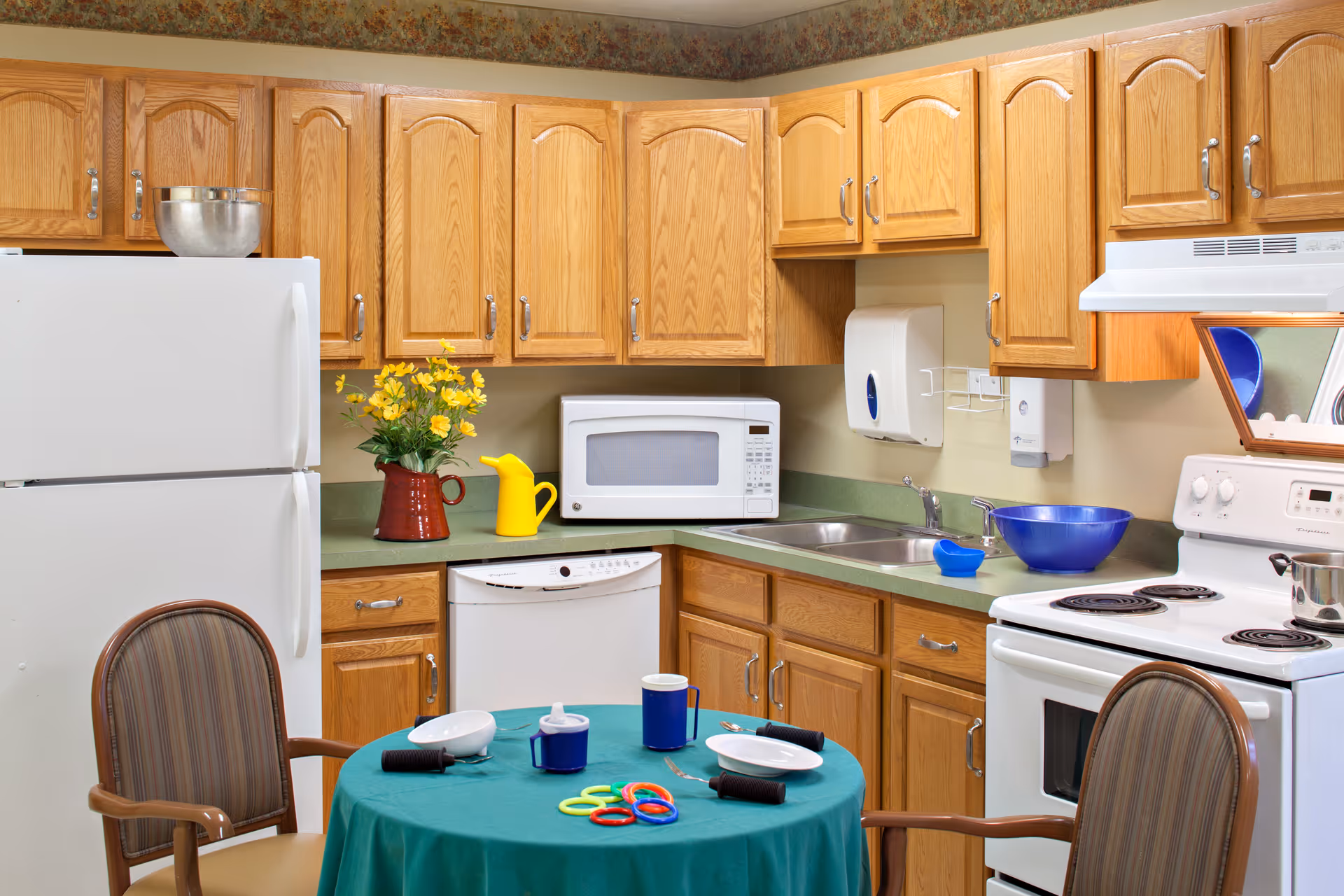 A kitchen area with wooden cabinets, a white refrigerator, microwave, dishwasher, and stove. A round table covered with a green tablecloth is set with two plates, cups, utensils, and colorful rings. Two chairs with striped upholstery are positioned around the table. A vase with yellow flowers and a yellow watering can are on the countertop.