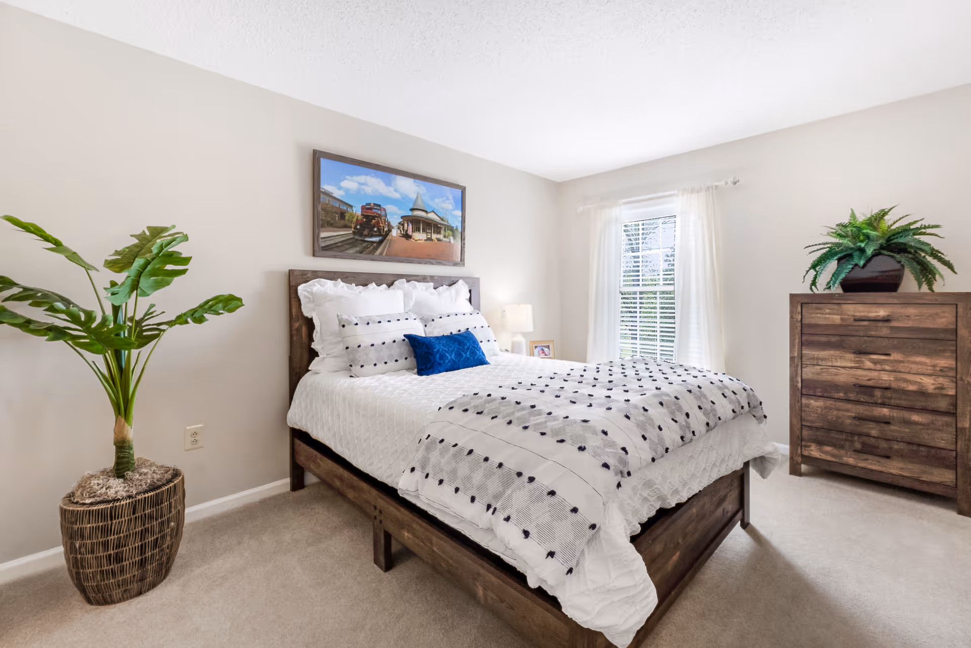 A bright and cozy bedroom with a wooden bed frame, white bedding with black polka dots, and a blue accent pillow. There is a large potted plant on the left side and a wooden dresser with a green plant on top on the right side. A window with white curtains lets in natural light, and a framed picture of a train station hangs above the bed.
