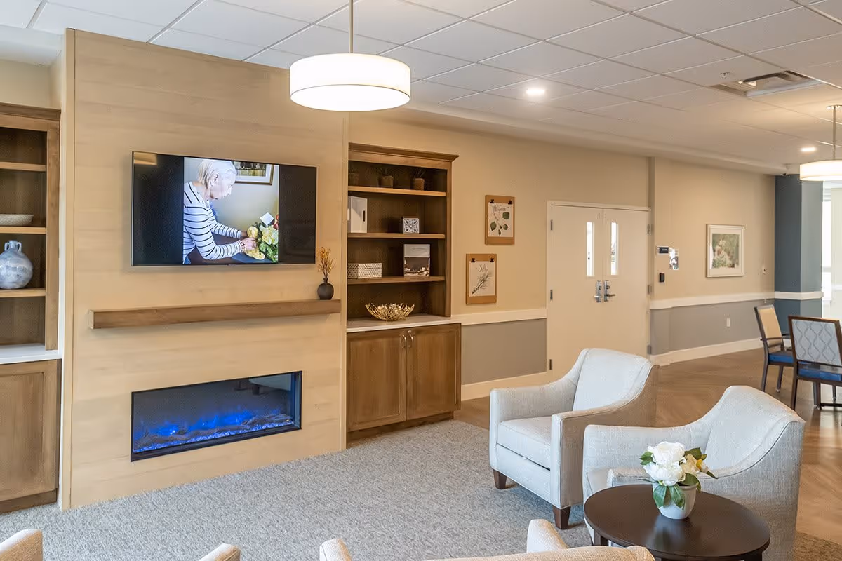 Comfortable common sitting area with two upholstered armchairs around a small table, a wall-mounted TV above a linear fireplace, built-in shelving, and dining chairs in the background.