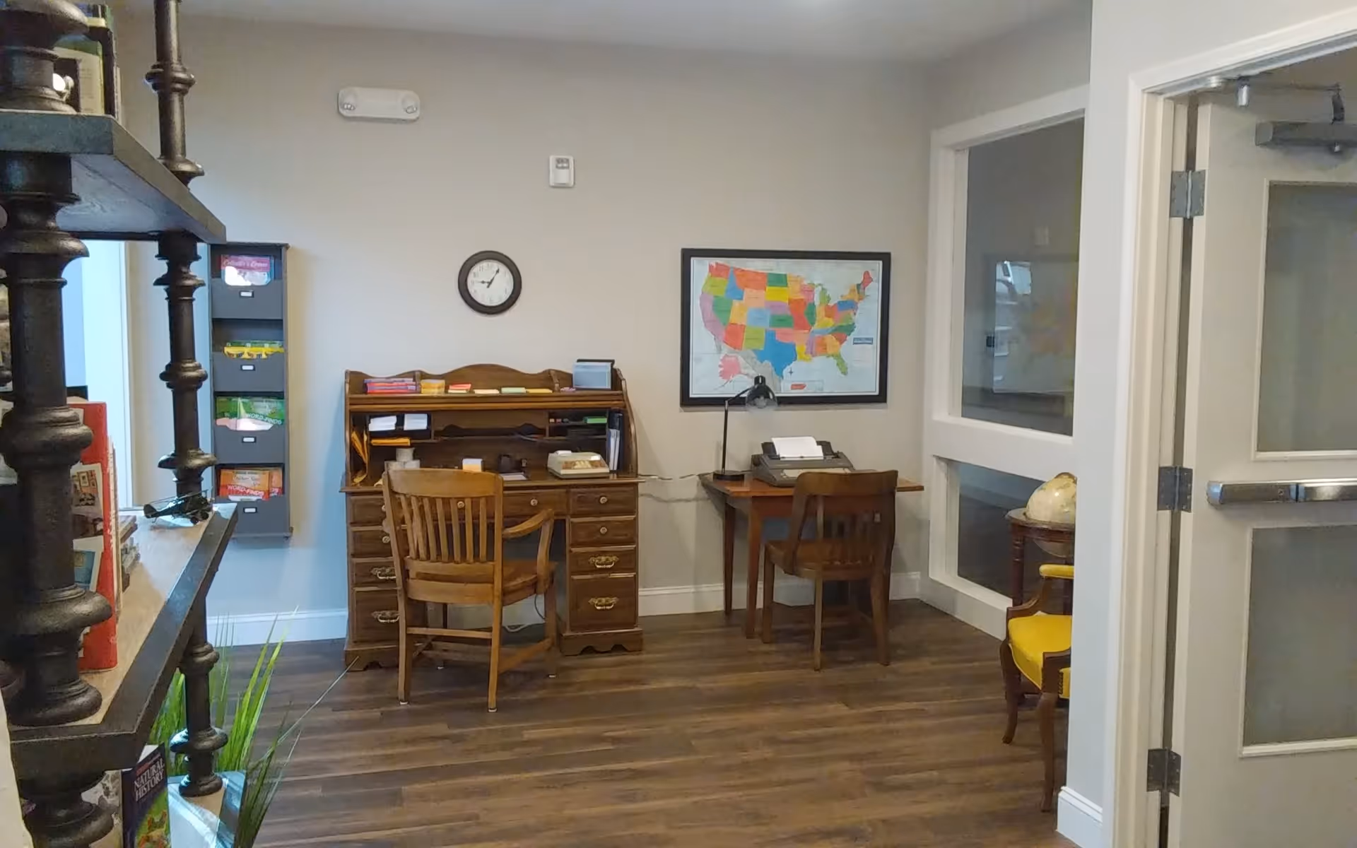 A small office or study area with two wooden desks and chairs. One desk has a vintage typewriter and a lamp, while the other has various office supplies. A framed map of the United States hangs on the wall above the desks, and a clock is mounted on the wall. To the left, there is a shelving unit with books and a wall-mounted organizer with colorful folders. The floor is wooden, and there is a door with glass panels on the right side.