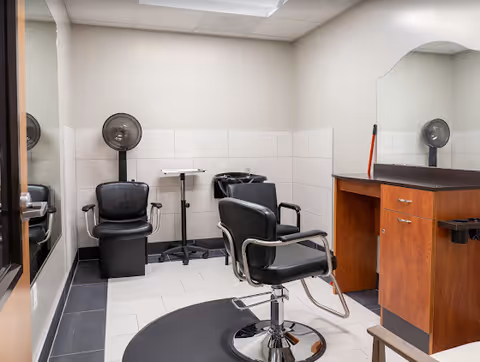 Small salon-style room with two black styling chairs, a shampoo sink, wall-mounted hair dryers, and a wooden styling station with mirror.