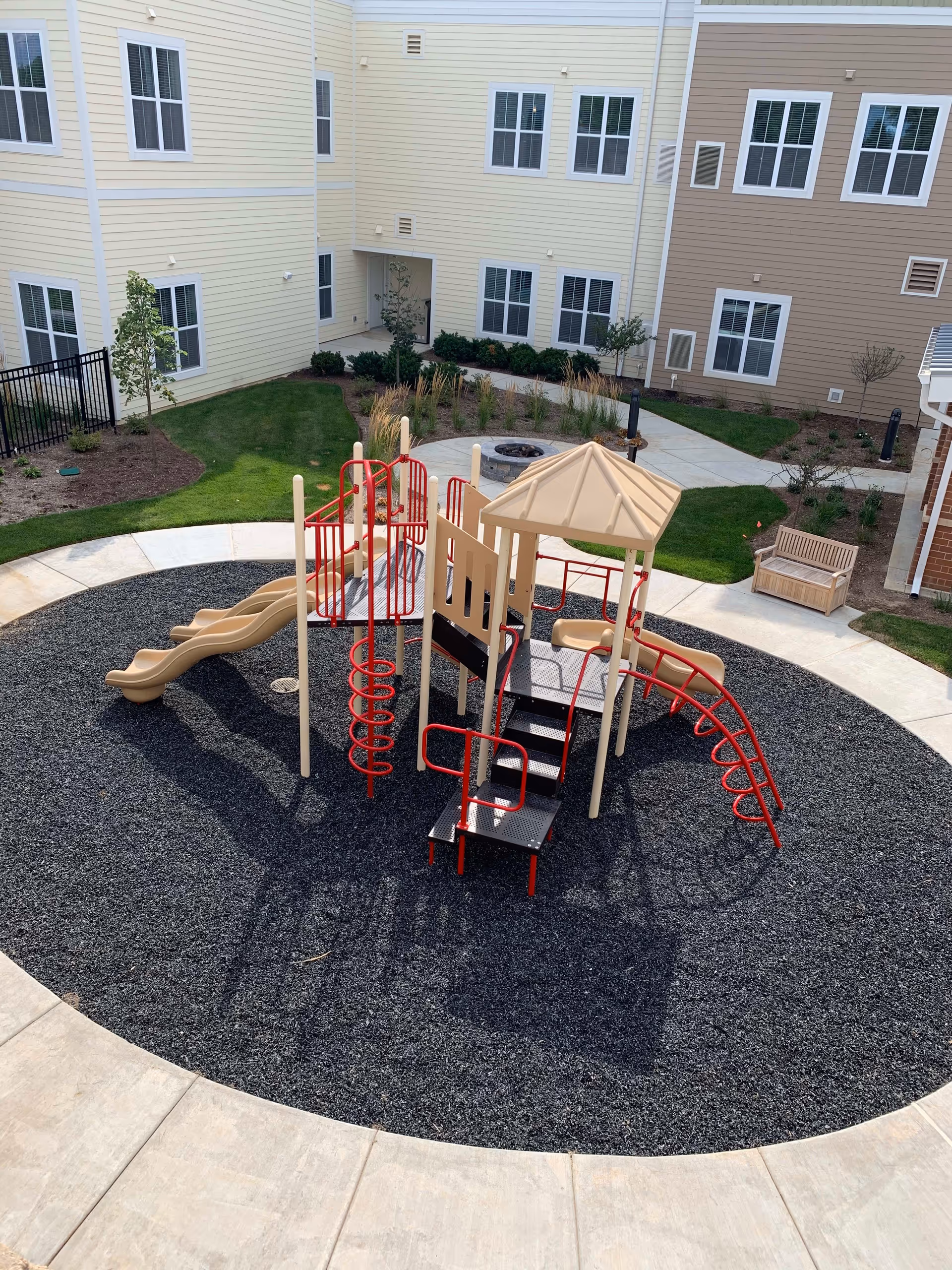 Outdoor playground area with beige and red play structures including slides, climbing bars, and steps, surrounded by black rubber mulch. The playground is situated in a courtyard with beige and light brown buildings, green grass, small trees, and a wooden bench nearby.
