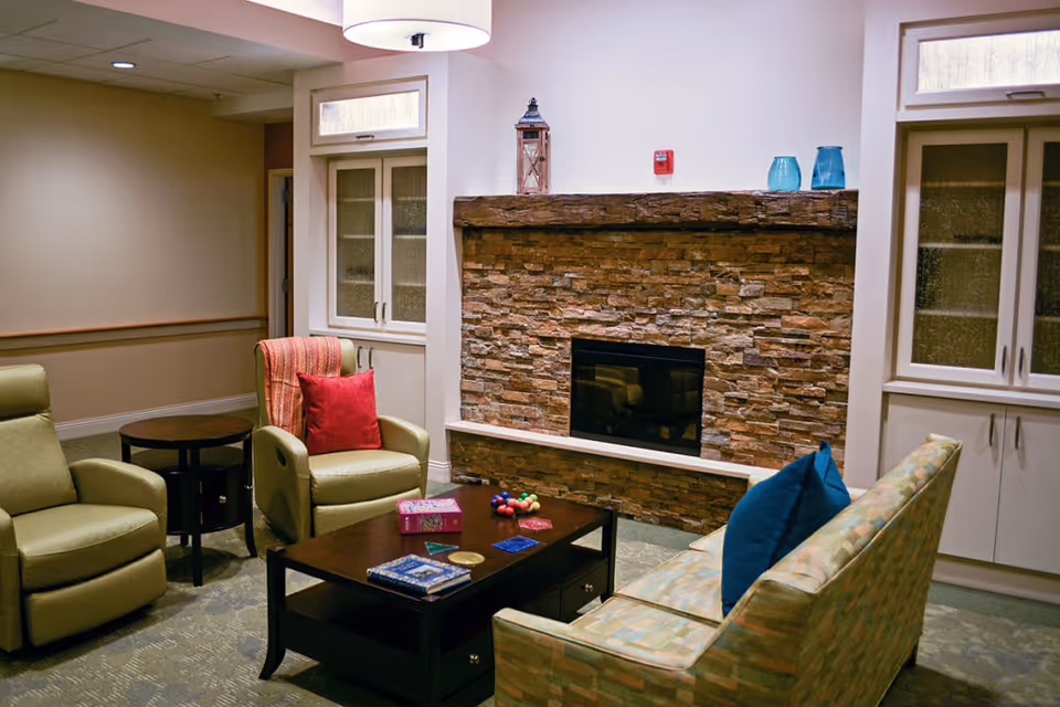 A cozy living room area with a stone fireplace, a wooden mantel decorated with a lantern and blue vases. The room has a patterned sofa with blue pillows, two armchairs including one with a red pillow, a dark wooden coffee table with board games and books, and built-in cabinets with glass doors on either side of the fireplace.