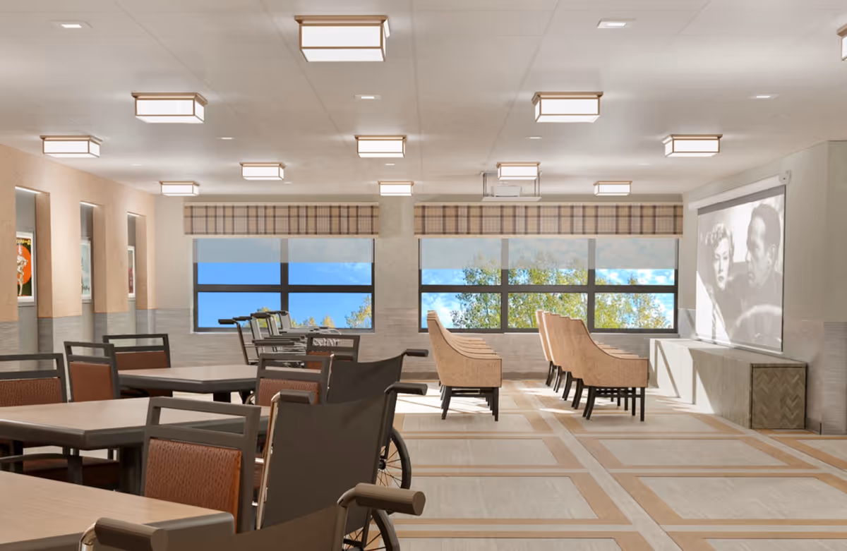 A spacious common area with multiple tables and chairs, including a wheelchair, facing a large screen showing a black-and-white movie. Large windows with plaid valances let in natural light and show trees outside. The ceiling has multiple square light fixtures.