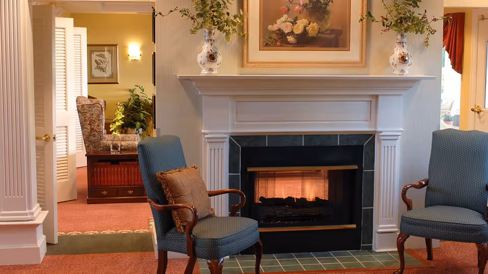 A cozy living room area featuring a lit fireplace with a white mantel, two upholstered armchairs with wooden arms and a brown cushion on one chair, floral vases on the mantel, and a framed floral painting above the fireplace. In the background, an open doorway reveals another room with a patterned armchair, a wooden chest, and some plants.