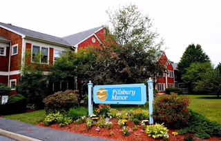 Exterior view of a red and white two-story building surrounded by trees and shrubs with a blue sign in front that reads 'Pillsbury Manor'. The area around the sign is landscaped with flowers and mulch.