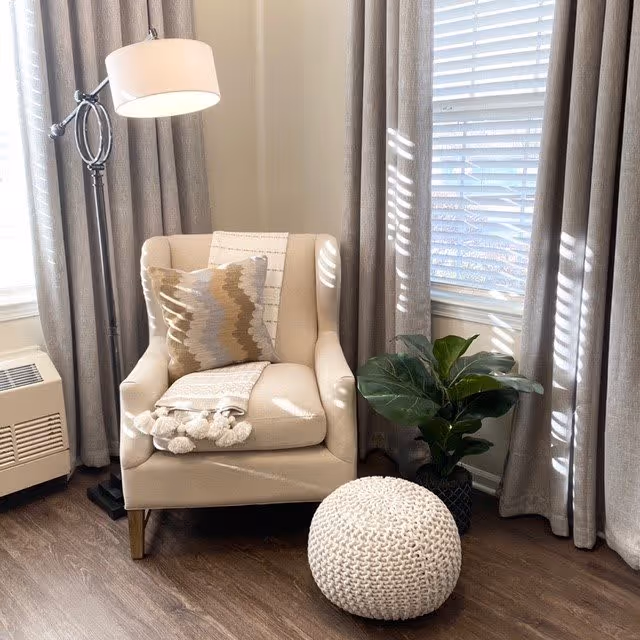 A cozy corner with a beige armchair adorned with a patterned pillow and a white throw blanket with pom-poms. Next to the chair is a white knitted pouf on a wooden floor, a green potted plant, and a tall floor lamp with a white lampshade. The background features beige curtains and a window with white blinds.