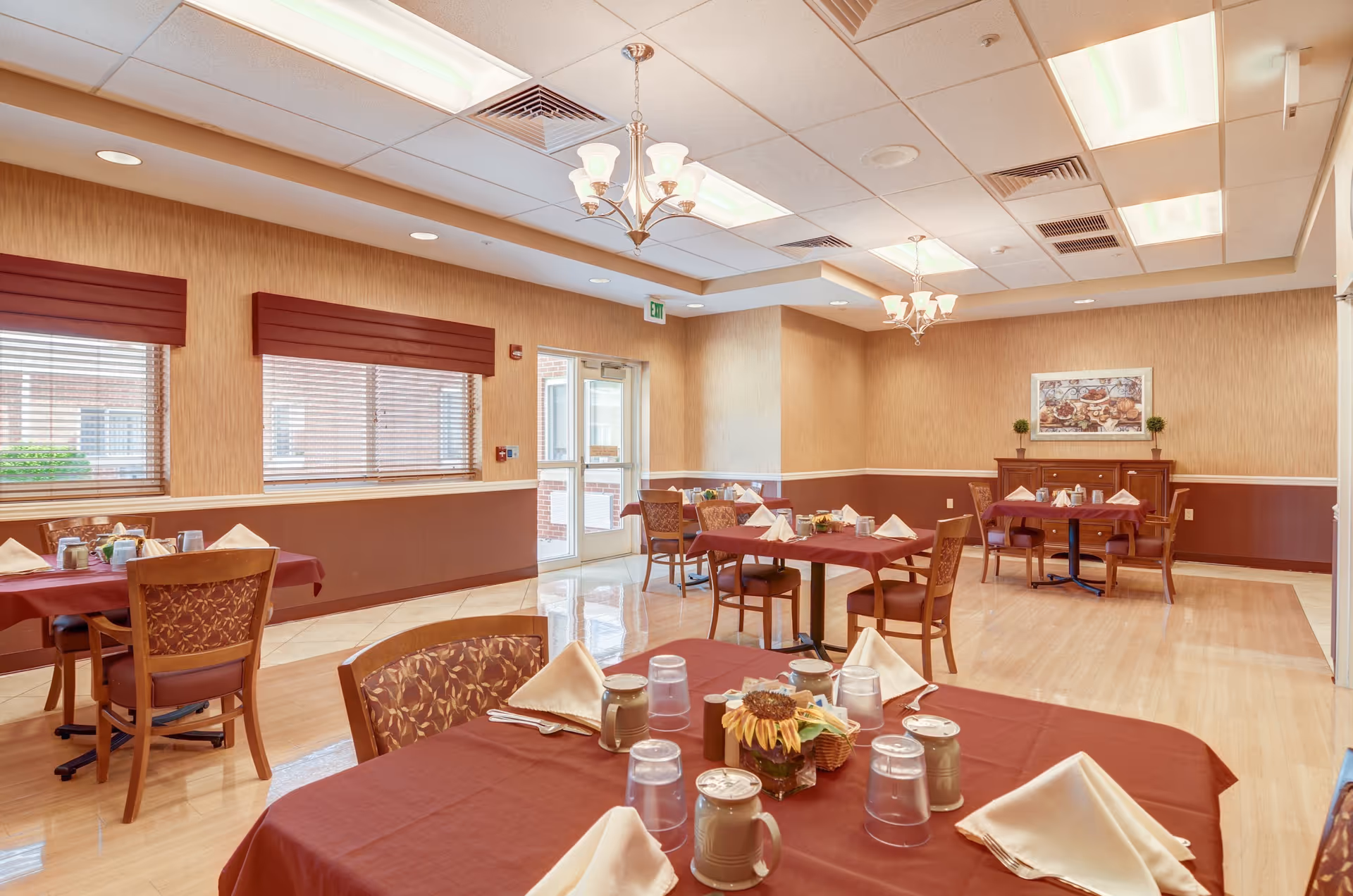 A dining room with several tables covered in maroon tablecloths, each set with beige napkins, cups, and glasses. The room has wooden chairs with patterned upholstery, beige walls with a maroon lower half, and windows with maroon valances. Two chandeliers hang from the ceiling, and a framed picture and small plants are on a wooden sideboard against the far wall.