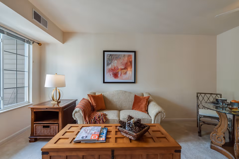 A cozy living room with a beige loveseat adorned with two orange pillows and a striped throw blanket. A wooden coffee table in front holds a basket with pinecones and some magazines. To the left, there is a wooden side table with a modern lamp, and a large window with blinds. On the right, a glass-top dining table with a cushioned chair is partially visible. A framed abstract painting hangs on the wall above the loveseat.