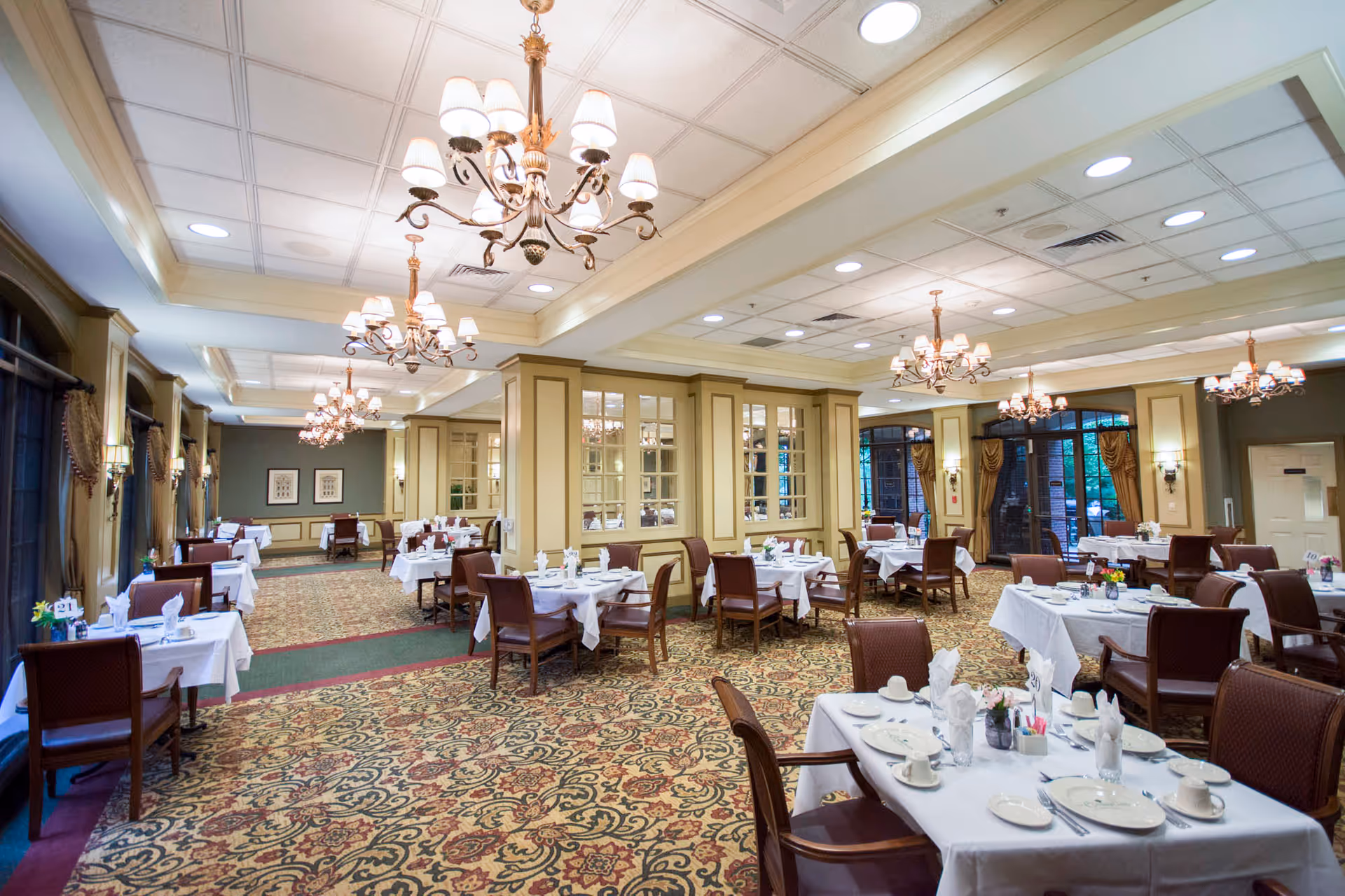 A spacious, elegant dining room with multiple tables covered in white tablecloths, set with plates, cups, and napkins. The room features patterned carpet, large windows with curtains, and ornate chandeliers hanging from the ceiling.
