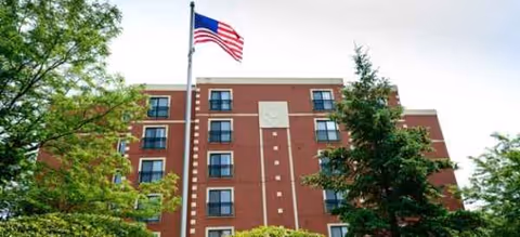 Brick senior living building with flagpole, American flag, and surrounding green trees.