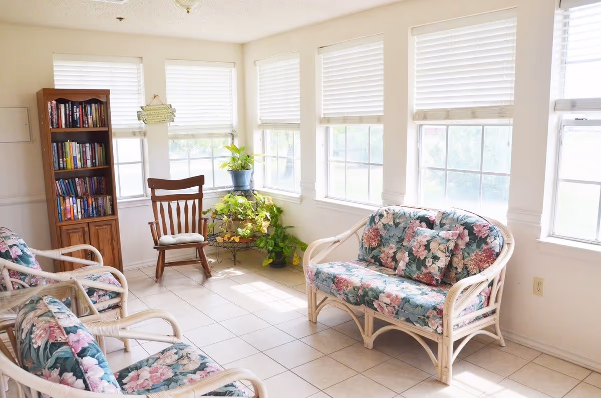 A bright and cozy sitting area with floral cushioned wicker chairs and a loveseat, a wooden rocking chair, a bookshelf filled with books, and several potted plants near large windows with white blinds.