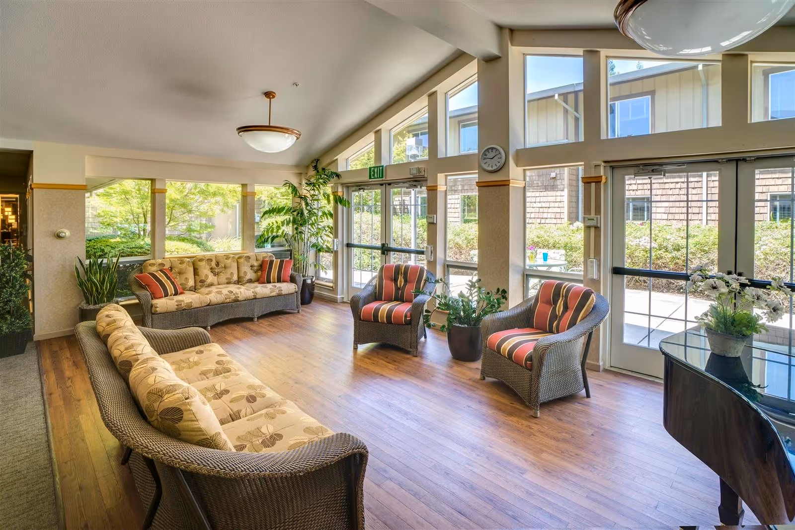 A bright and spacious living room area with large windows allowing natural light to fill the space. The room features two cushioned wicker sofas with floral patterns, two cushioned wicker armchairs with striped upholstery, several potted plants, a wall clock, and a glass-top table. Outside the windows, greenery and a building exterior are visible.