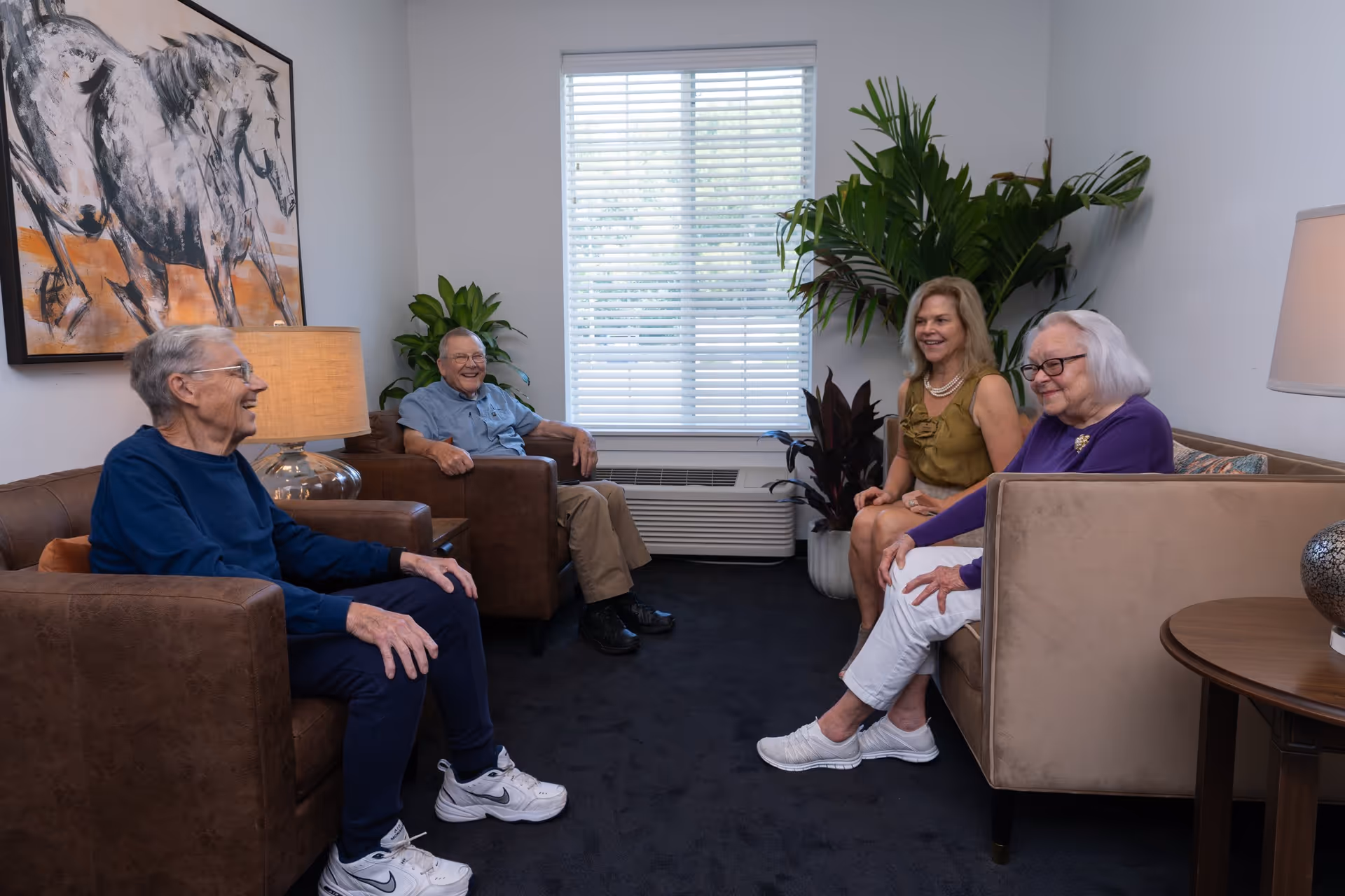 Four elderly people sitting and smiling in a well-lit living room with brown leather chairs and a beige sofa. There is a large window with blinds, green plants, a table lamp, and a painting of horses on the wall.