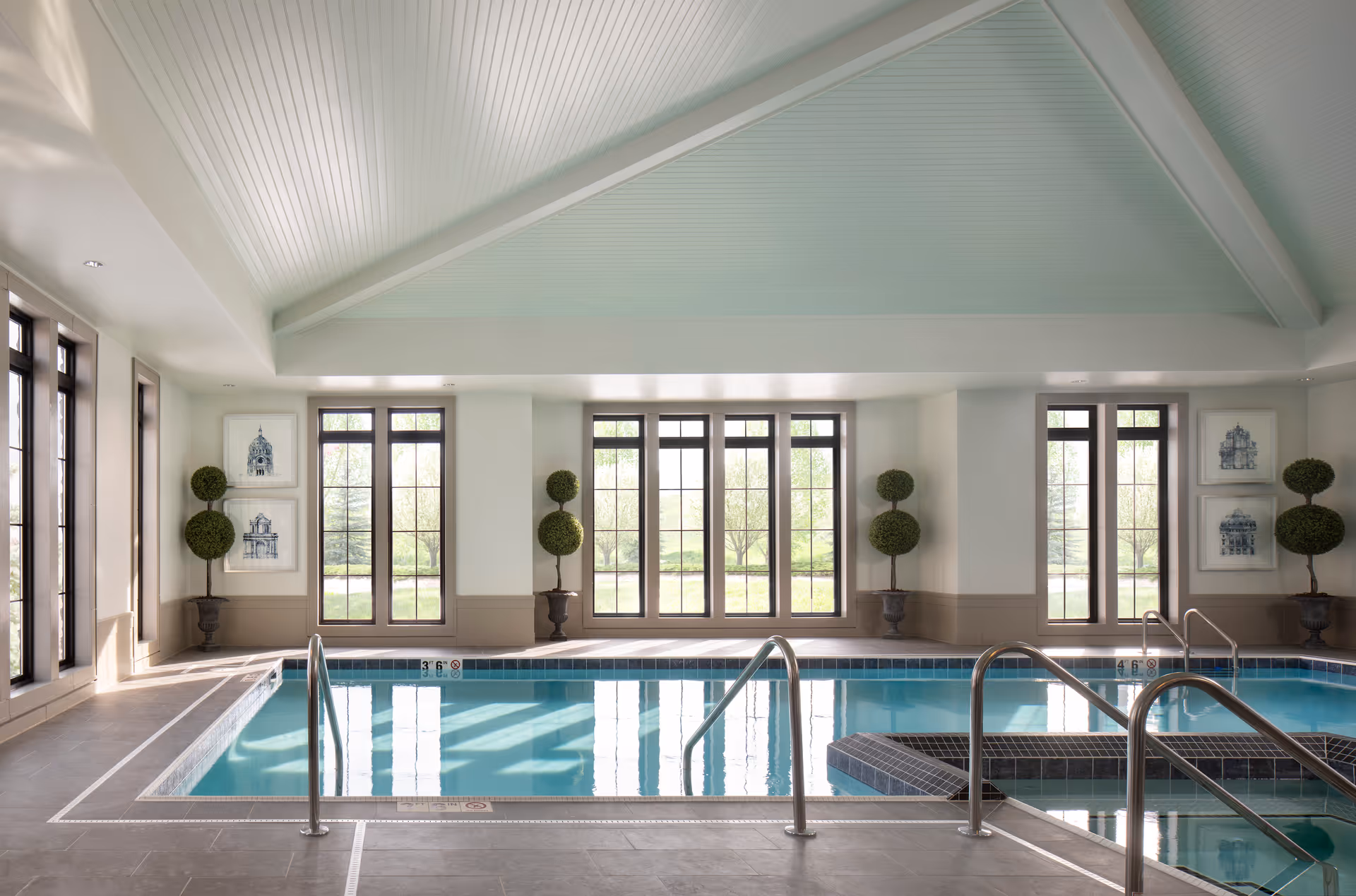 Indoor swimming pool area with large windows letting in natural light. The pool has metal handrails and steps for easy access. The room features a high, vaulted ceiling with white paneling, potted topiary plants, and framed artwork on the walls.