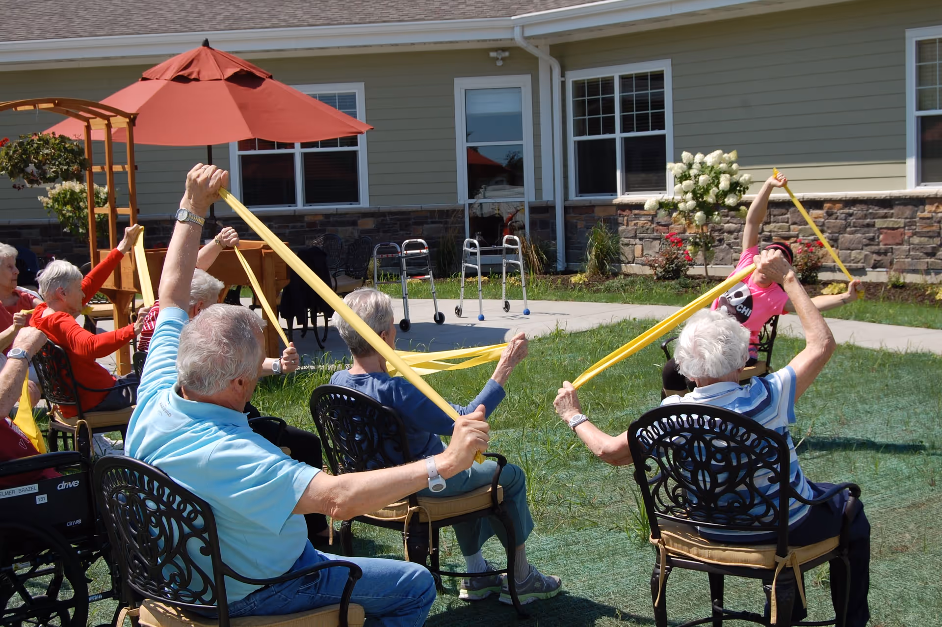 A group of elderly people seated outdoors in a garden area participating in a seated exercise session using yellow resistance bands. They are arranged in a semi-circle with a staff member leading the exercise. The background shows a building with windows, a red umbrella, and some walkers parked nearby.