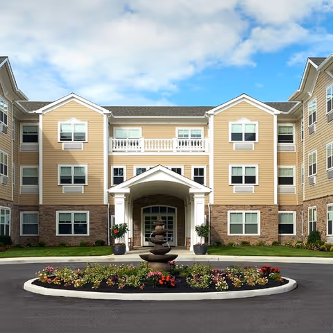 Front exterior view of a three-story senior living facility building with beige siding and stone accents. The entrance features a covered portico with white columns, and there is a circular driveway with a landscaped island in the center containing a tiered water fountain and colorful flowers. The sky is partly cloudy.