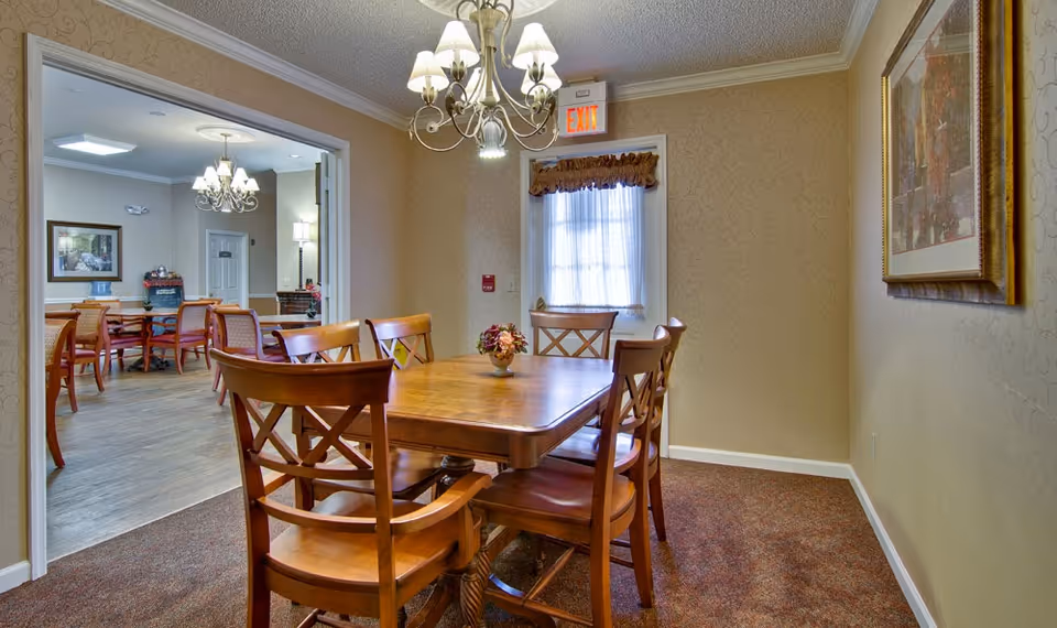 A cozy dining area in a senior living facility featuring a wooden table with six matching chairs. A small floral centerpiece decorates the table. The room has beige patterned wallpaper, a framed painting on the right wall, and a window with a curtain and valance. A chandelier hangs above the table, and an exit sign is visible above the window. Through an open doorway, another dining area with additional tables and chairs and a beverage station can be seen.