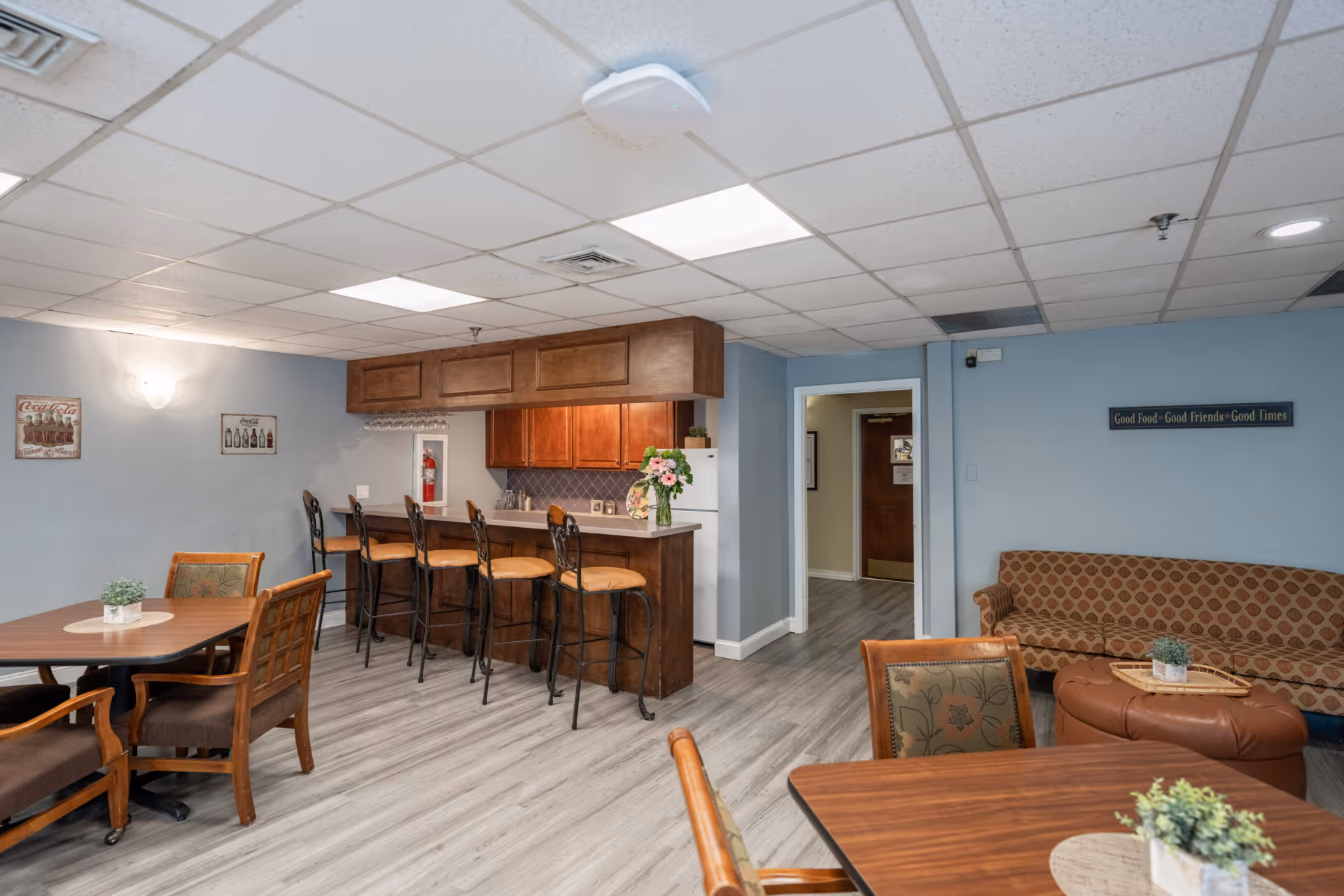 A communal dining and lounge area in a senior living facility featuring wooden tables and chairs, a kitchen counter with four high chairs, a patterned couch, and wall decorations including a sign that reads 'Good Food - Good Friends - Good Times'.