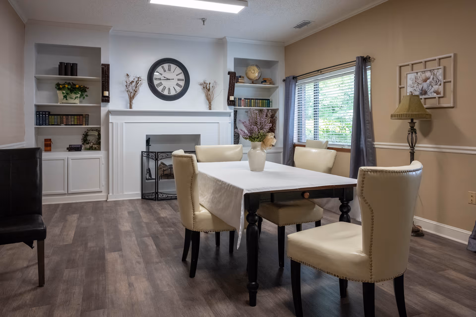 A cozy dining room with a rectangular table covered by a white tablecloth and surrounded by four cream-colored upholstered chairs. The room features a fireplace with a large round clock above it, built-in shelves with books and decorative items, a window with gray curtains, a floor lamp, and a framed floral picture on the wall.