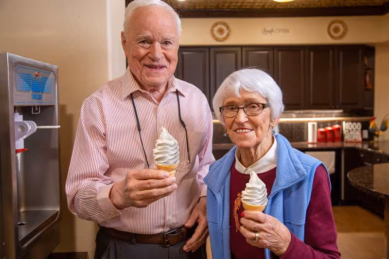 An elderly man and woman standing indoors in a kitchen area, each holding a soft serve ice cream cone and smiling at the camera. The man is wearing a striped shirt and the woman is wearing glasses, a maroon top, and a blue vest. Behind them are dark kitchen cabinets and a countertop with various items.