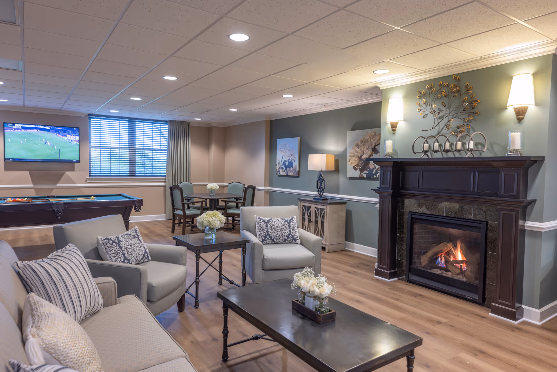 A cozy living room area in an assisted living facility featuring a lit fireplace with decorative wall art above it, a flat-screen TV mounted on the wall showing a sports game, a pool table, a round table with chairs, and comfortable seating including a sofa and armchairs with patterned cushions. The room has wooden flooring, soft lighting from wall sconces and a table lamp, and large windows with blinds.
