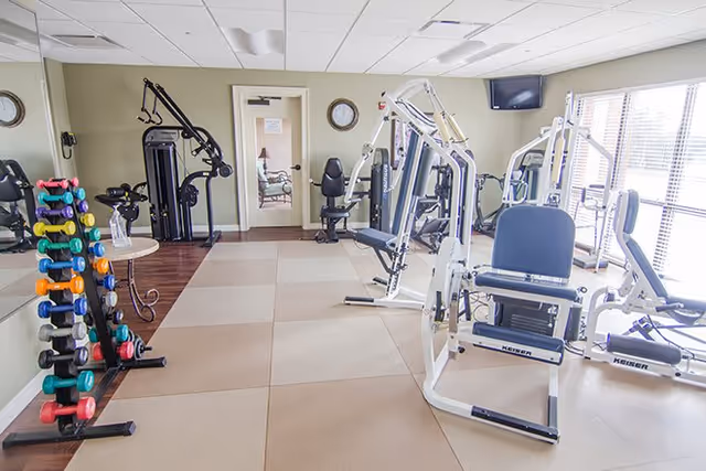 A bright fitness room with various exercise equipment including weight machines, a rack of colorful dumbbells, and a stationary bike. The room has large windows letting in natural light, a wall mirror, and a wall-mounted TV.