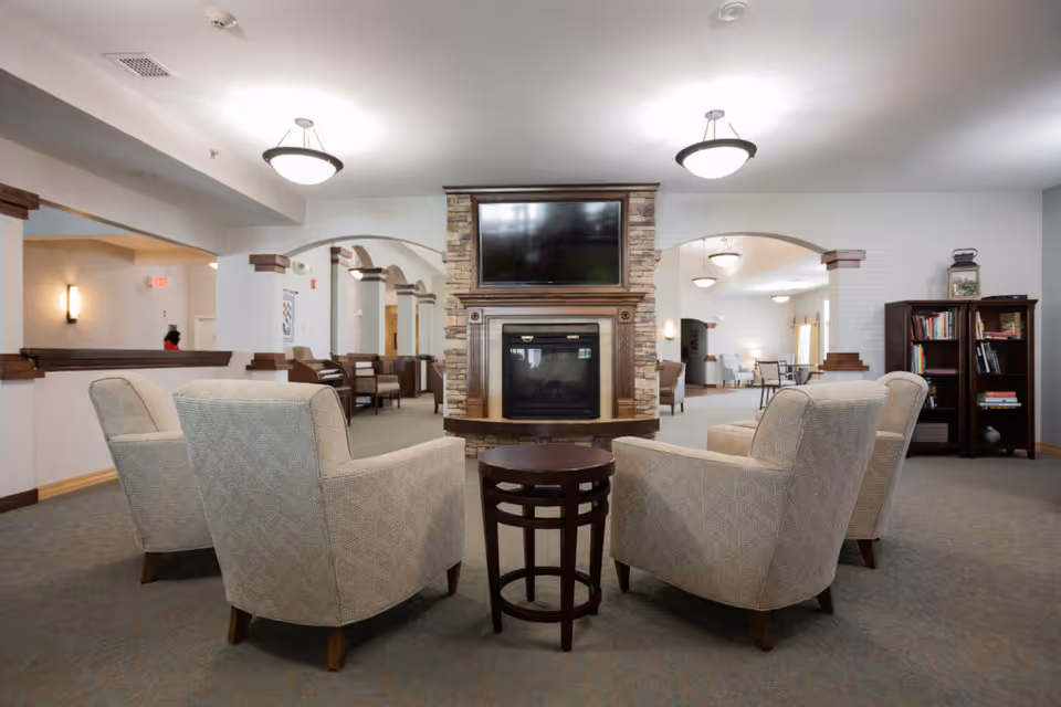 A cozy common area in a senior living facility featuring four beige upholstered armchairs arranged around a small round wooden table. In the center background, there is a stone fireplace with a mounted flat-screen TV above it. The room has soft lighting with ceiling fixtures and wall sconces, and there are bookshelves and additional seating visible in the background.