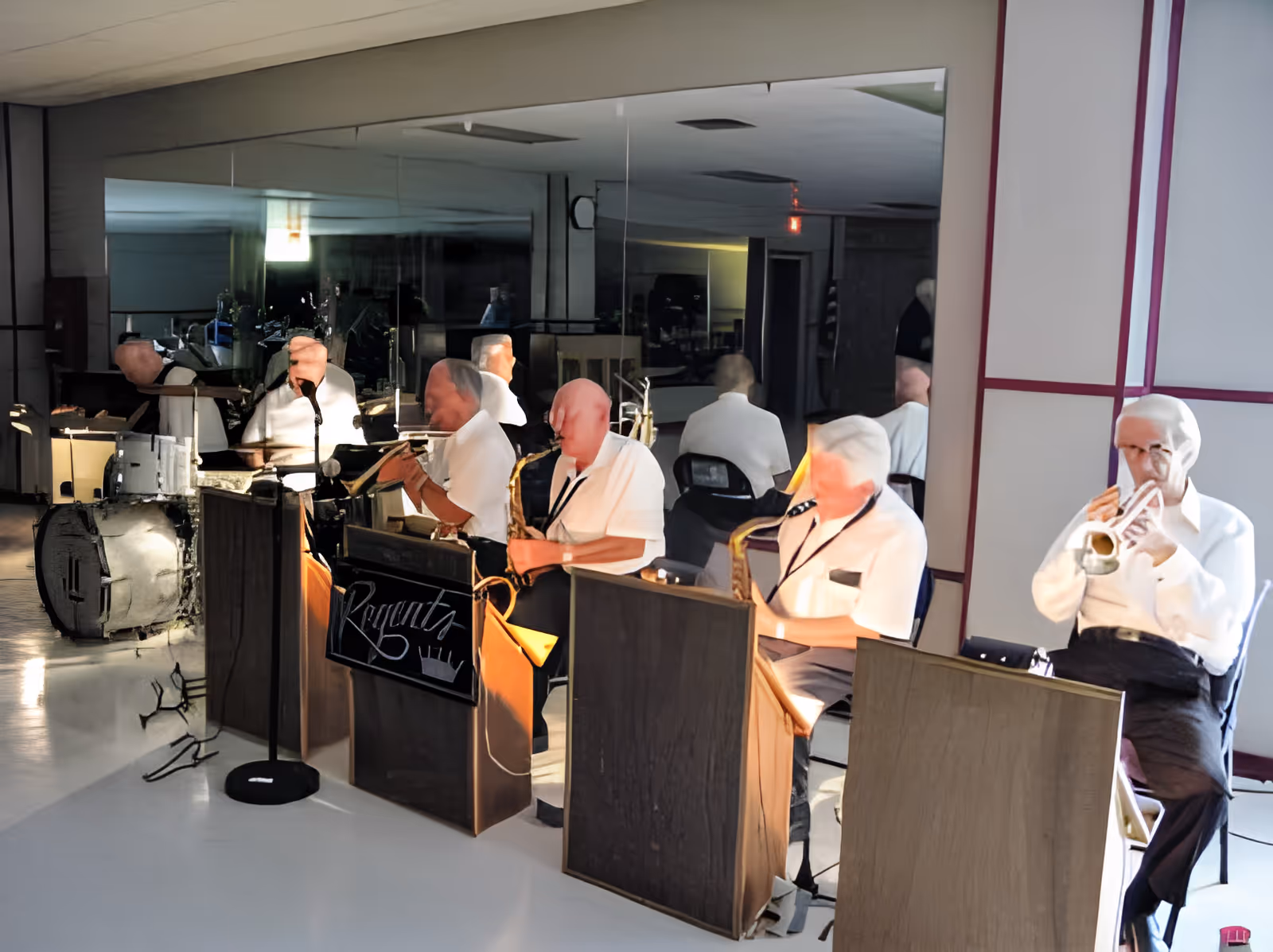 A group of elderly musicians playing instruments including saxophones, trumpet, and drums in an indoor setting with a large mirror behind them reflecting their images.
