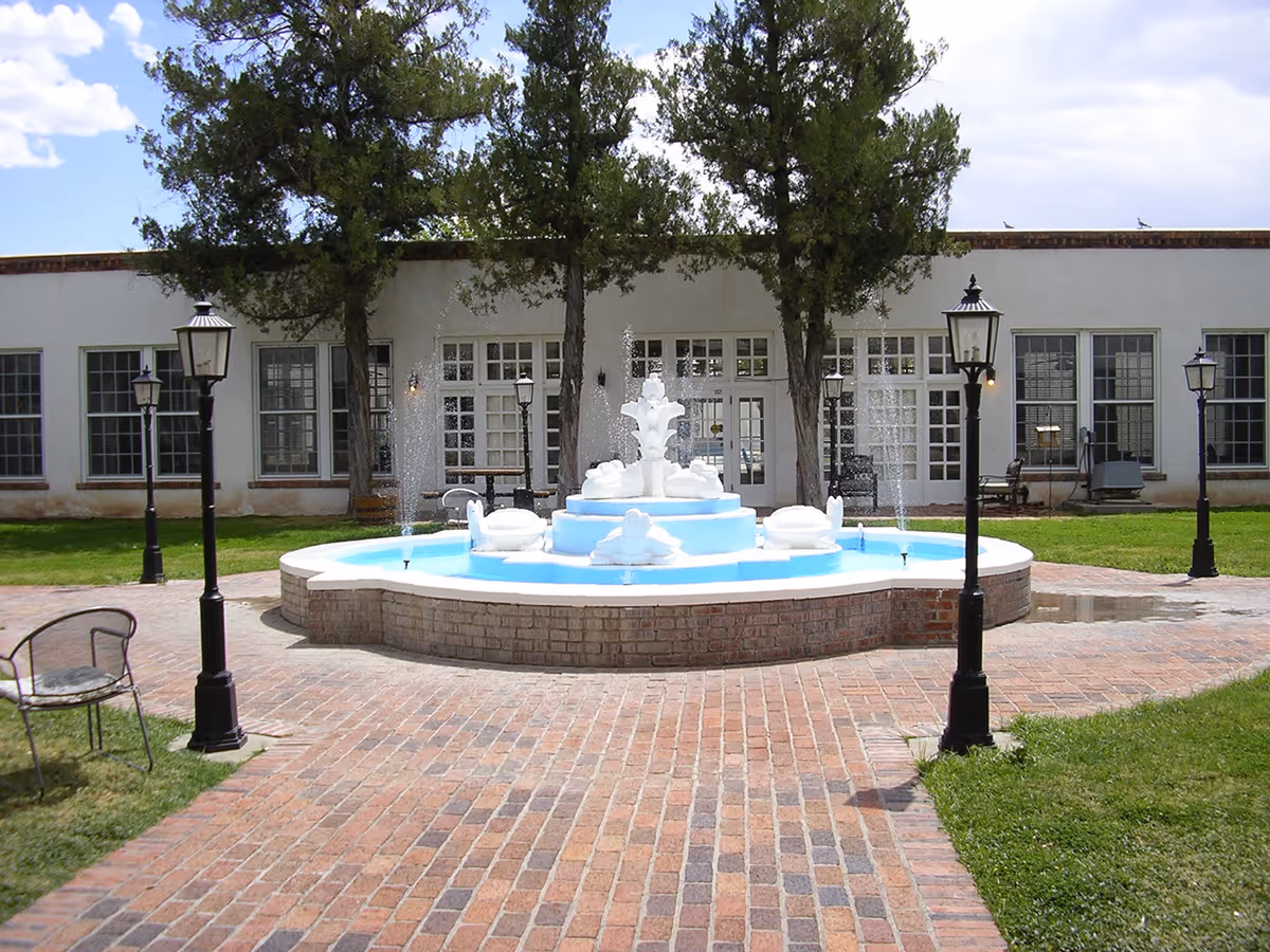 Brick courtyard with a central multi-tiered white fountain surrounded by lamp posts in front of a single-story building with large windows.
