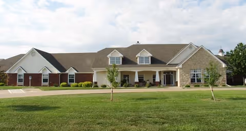 Exterior view of a single-story senior living facility building with a combination of brick and stone facade, multiple windows, and a covered entrance. The building is surrounded by a well-maintained lawn with a few small trees and a paved driveway in front.