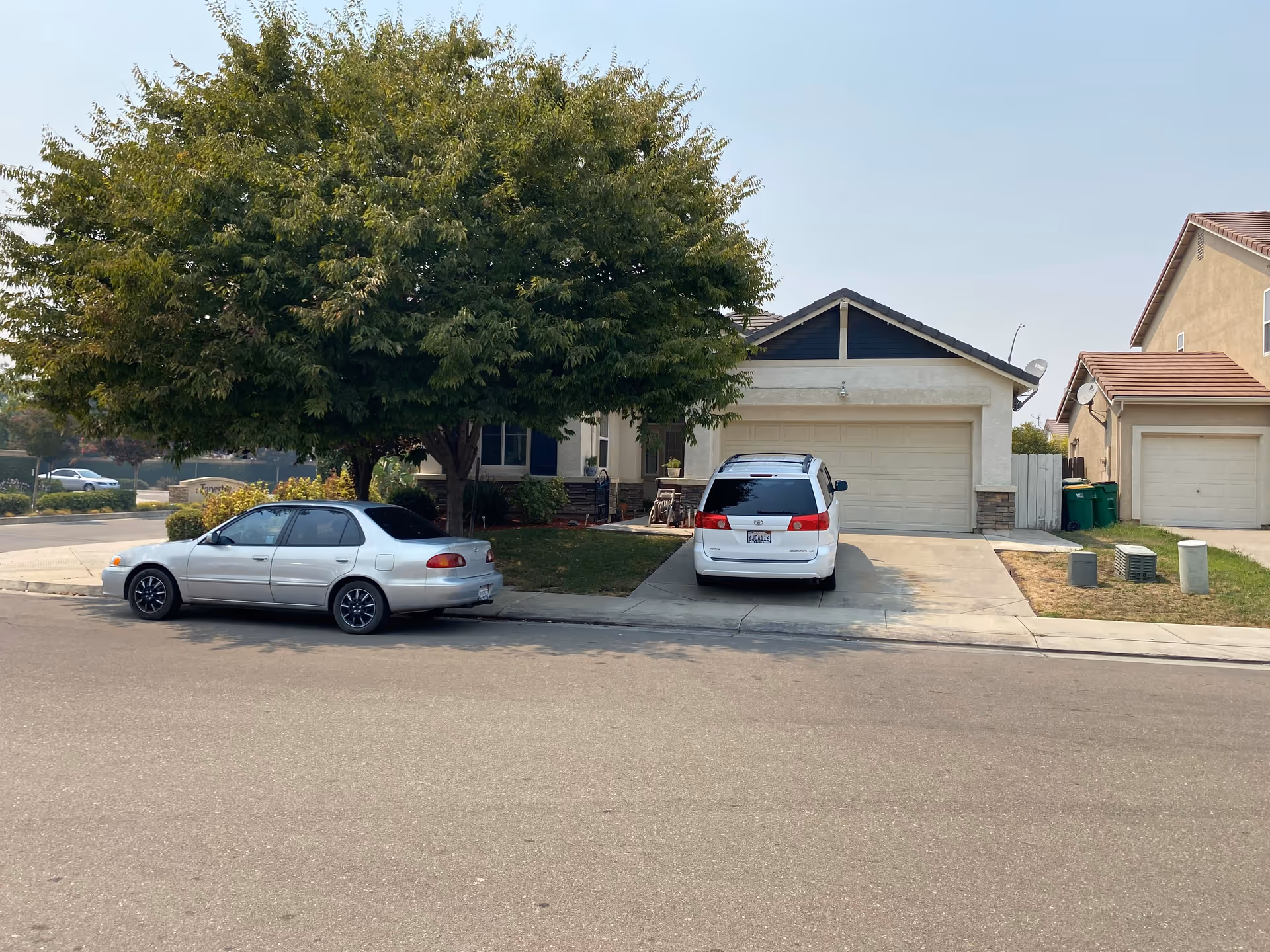 Suburban house facade with large tree, two parked cars, and a driveway.