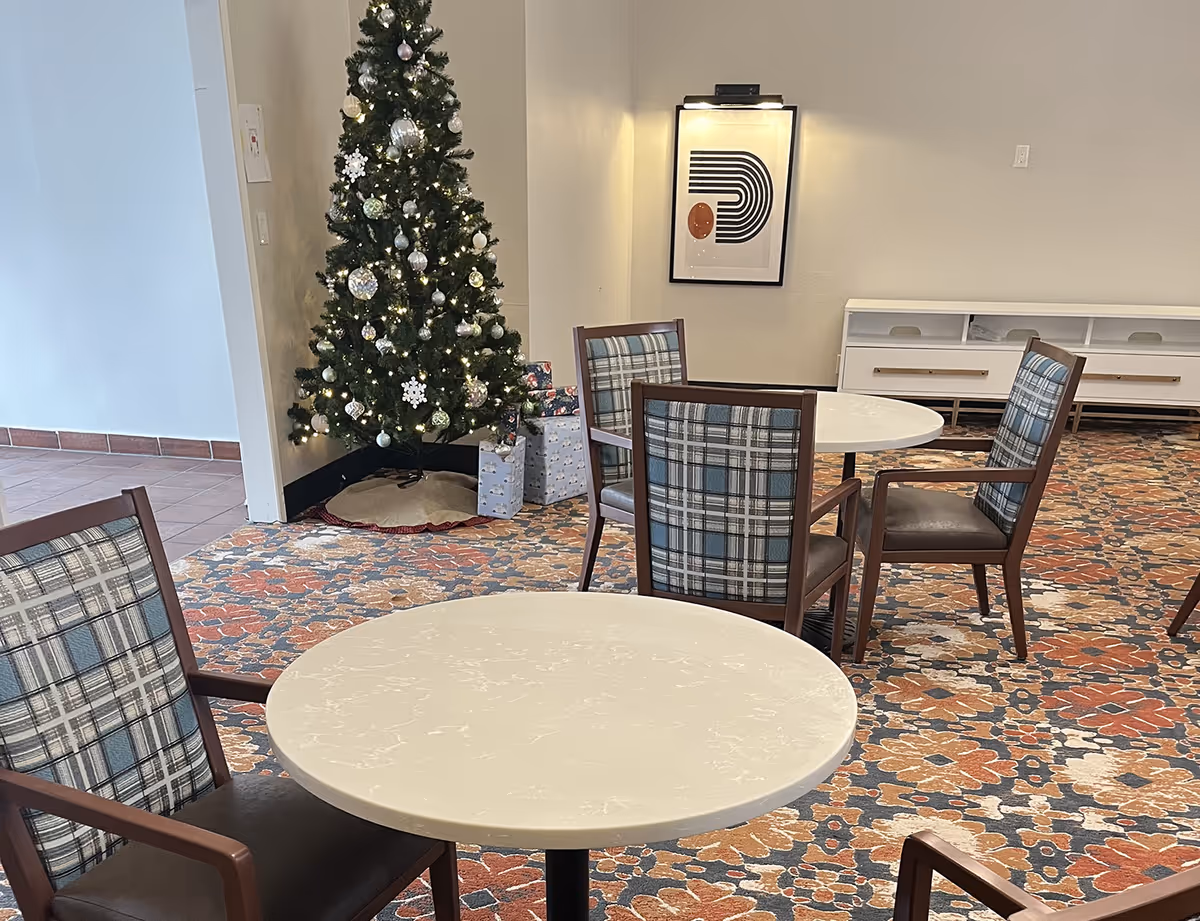 A common area with round tables and chairs featuring plaid upholstery. A decorated Christmas tree with lights and ornaments stands in the corner with wrapped presents underneath. A framed abstract artwork is mounted on the wall above a white storage cabinet. The floor is covered with a floral patterned carpet.